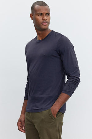 A man wearing a long-sleeved navy blue SKEETER TEE by Velvet by Graham & Spencer and olive green pants stands with one hand in his pocket, looking to the side against a plain background. His choice of attire is a versatile wardrobe addition, effortlessly blending comfort and style.