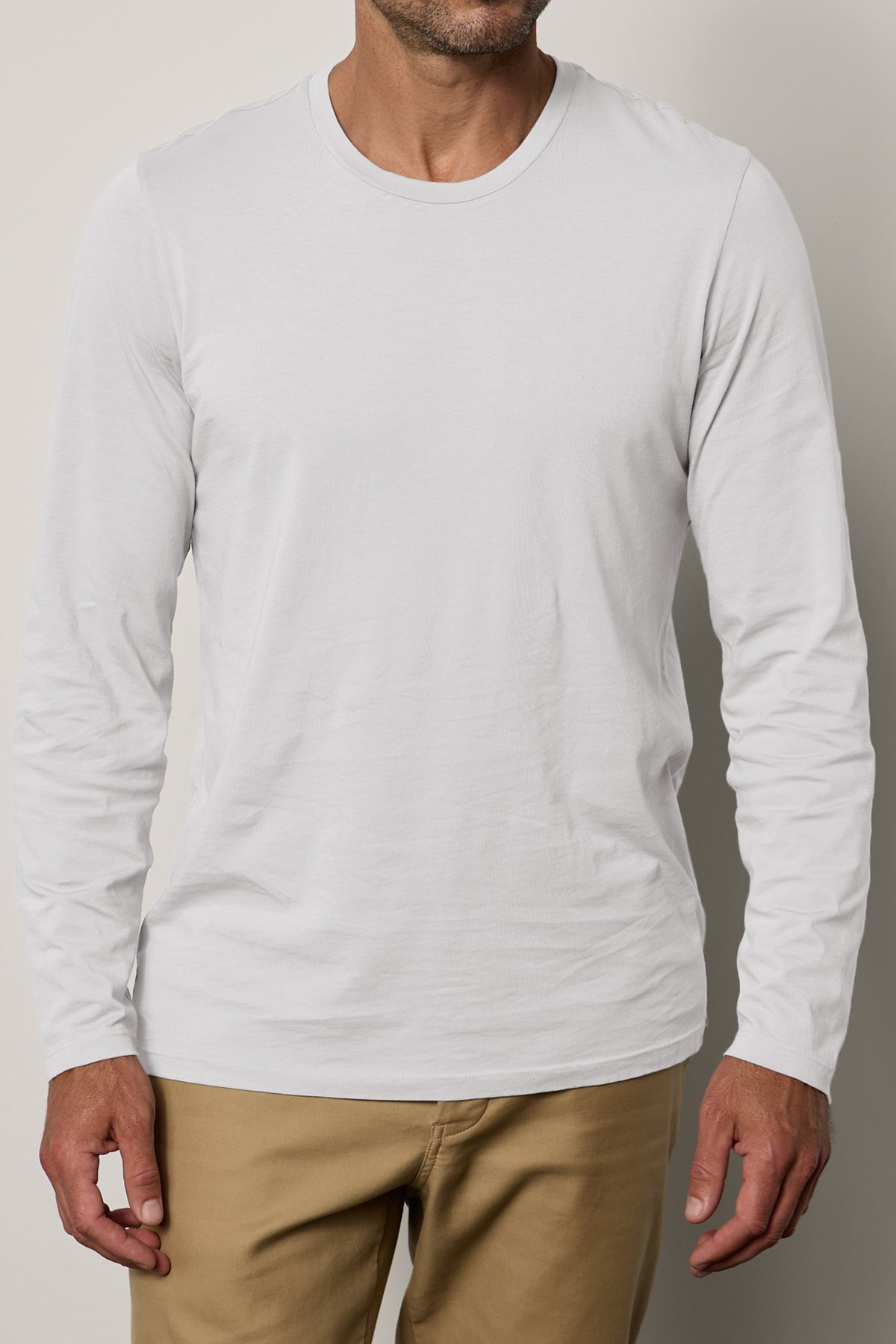   A person wearing a classic crew neckline Velvet by Graham & Spencer SKEETER TEE in plain white long-sleeve Pima cotton and beige pants stands against a neutral background. The image captures the upper body from the shoulders to mid-thigh. 