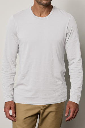 A person wearing a classic crew neckline Velvet by Graham & Spencer SKEETER TEE in plain white long-sleeve Pima cotton and beige pants stands against a neutral background. The image captures the upper body from the shoulders to mid-thigh.