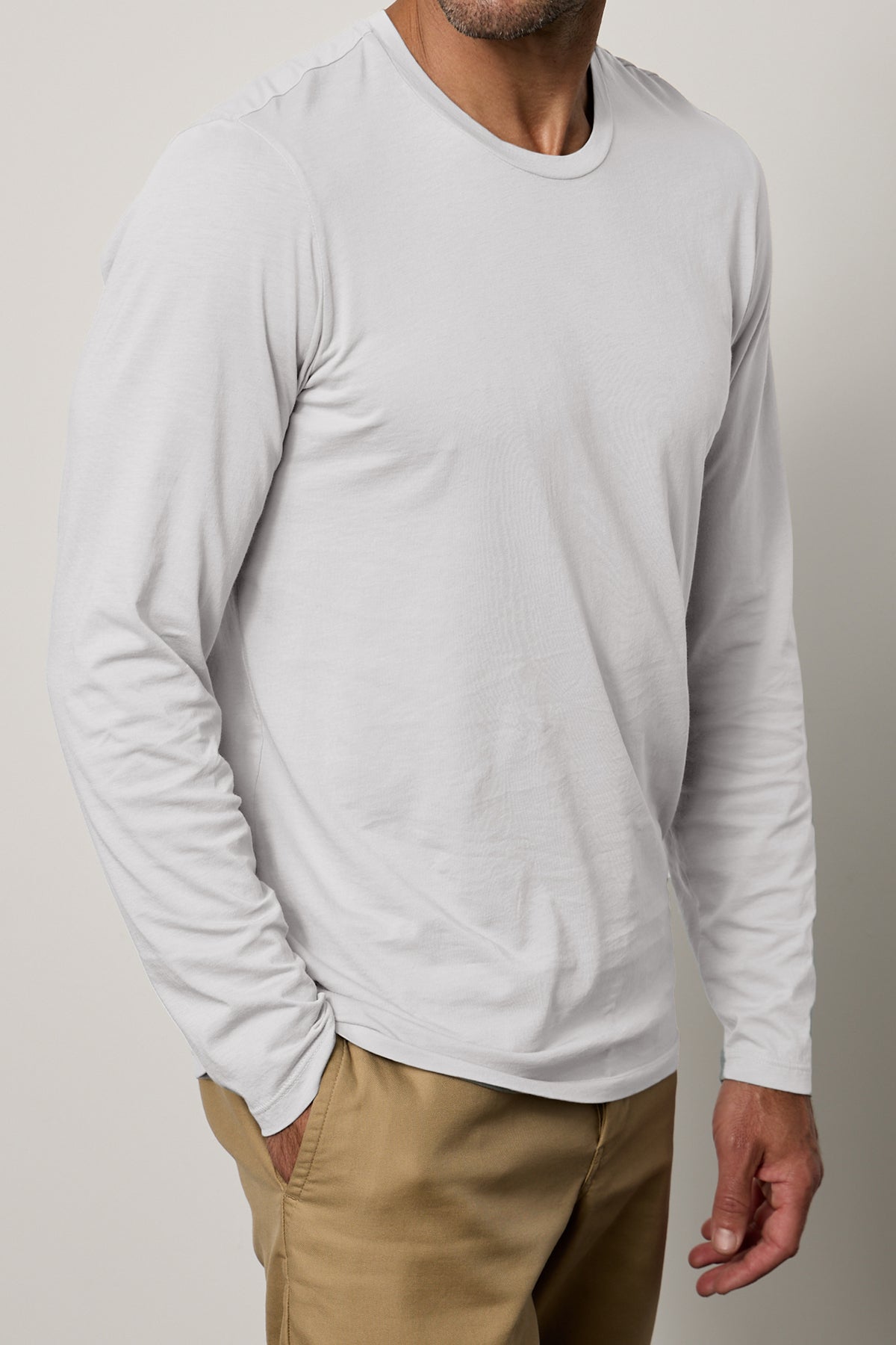   A person dressed in a plain, long-sleeved white SKEETER TEE by Velvet by Graham & Spencer with a classic crew neckline and beige pants, one hand casually in their pocket, stands against a neutral background. 