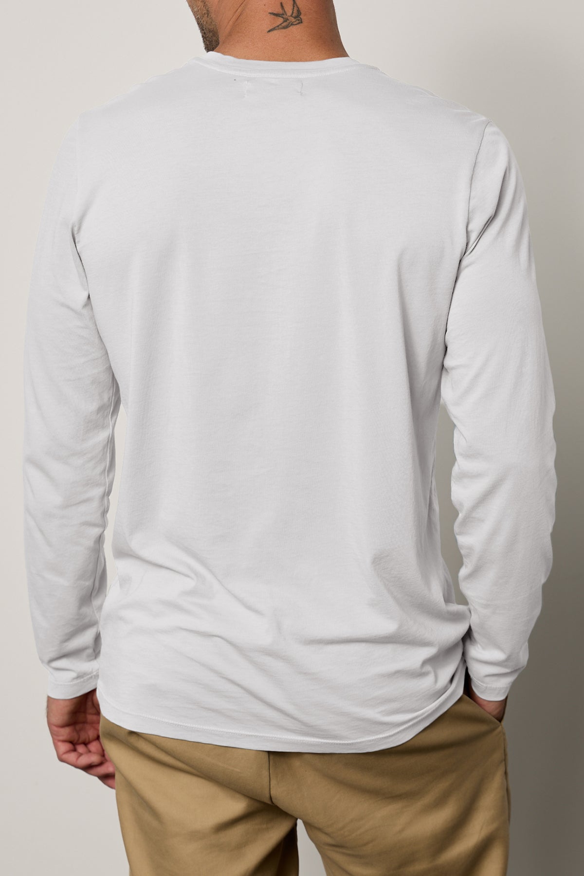   A person wearing a white, long-sleeved Velvet by Graham & Spencer Skeeter Tee and beige pants, shown from the back. The classic crew neckline complements the elegant simplicity, while the soft Pima cotton fabric enhances comfort. The person's neck features a small tattoo. 