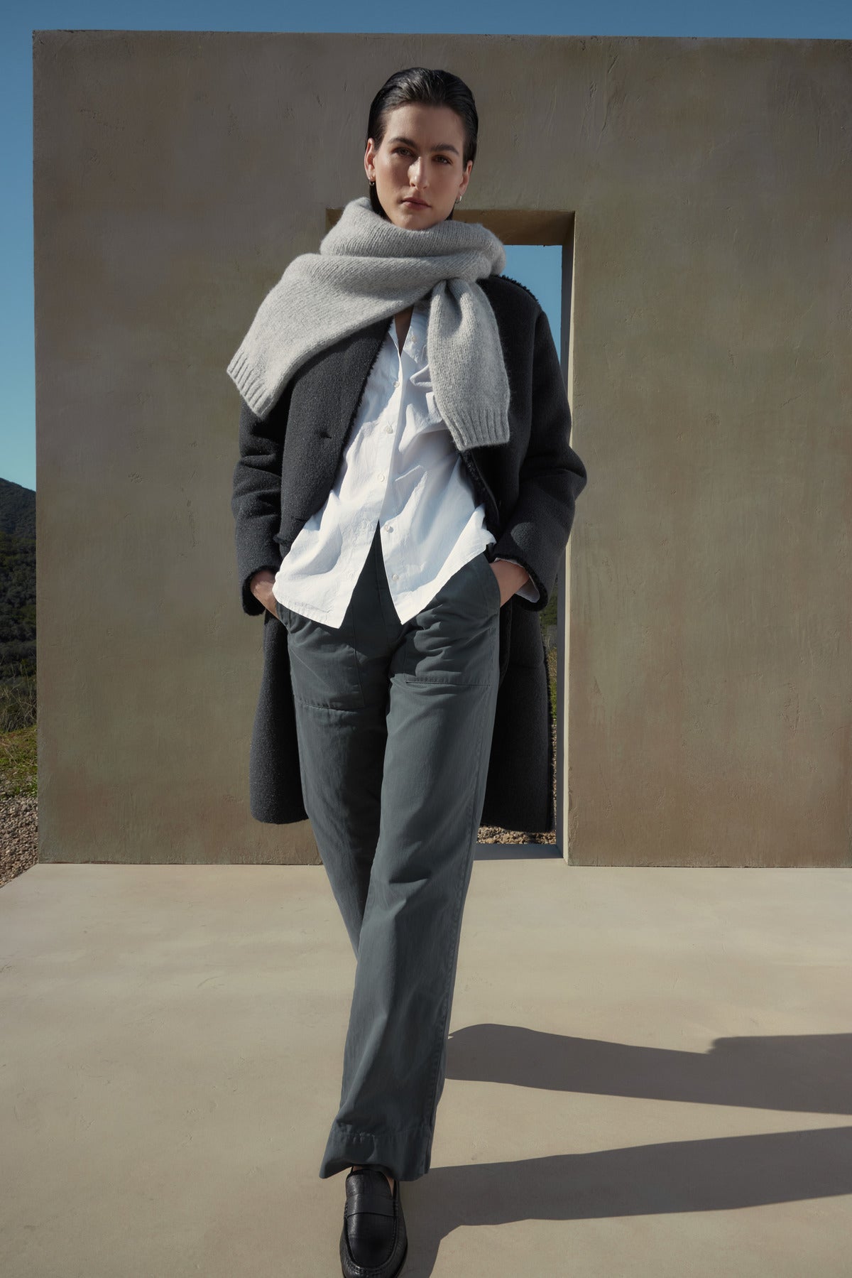   A person stands outdoors, wearing a dark coat, white shirt, cotton twill scarf, and the VENTURA PANT by Velvet by Jenny Graham. A concrete structure with a rectangular opening is behind them. 