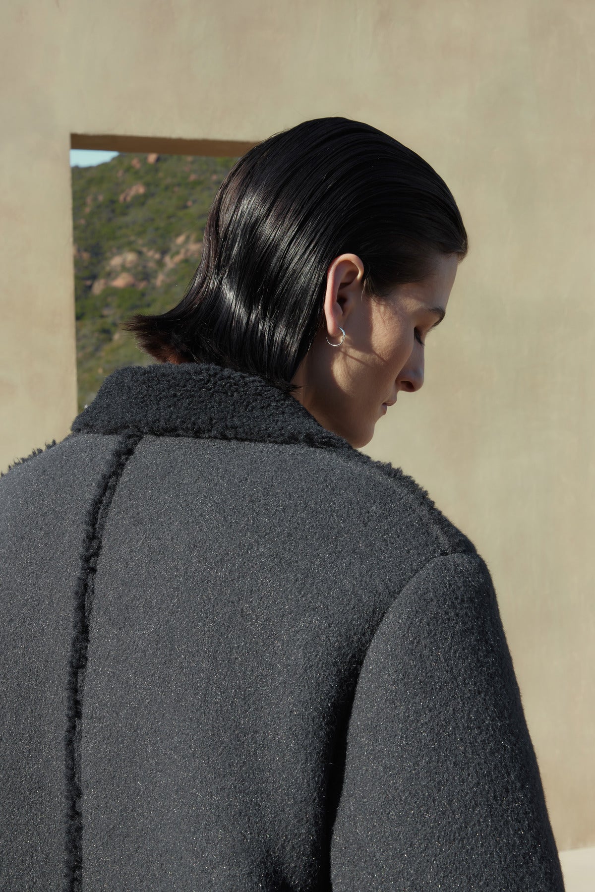 A person with dark, slicked-back hair is seen from behind, wearing the ROSEMEAD COAT by Velvet by Jenny Graham. The background features an outdoor setting with subtle landscape elements visible through an opening.-37666838610113