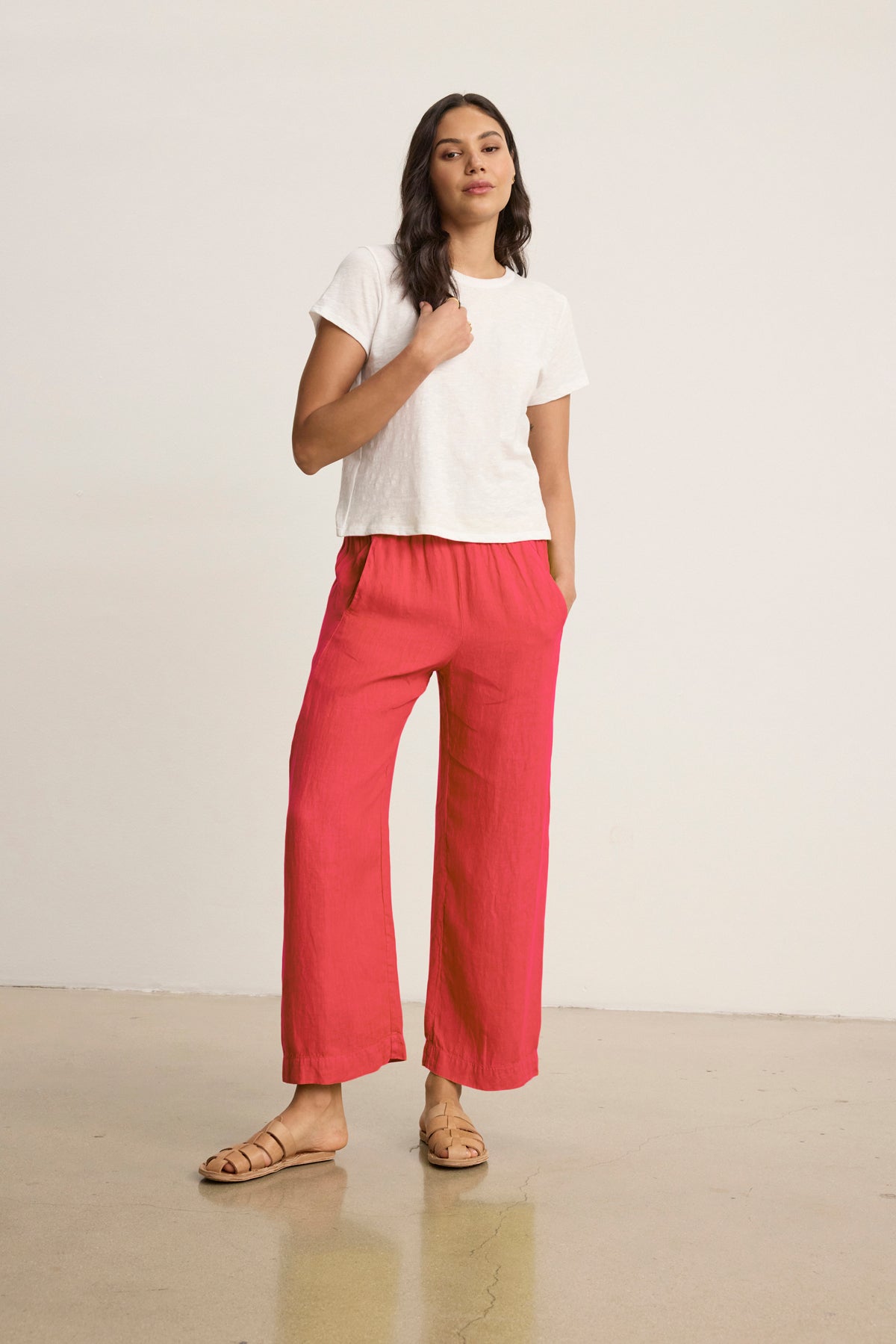   Someone wearing the LOLA LINEN PANT by Velvet by Graham & Spencer, styled with a white t-shirt, stands relaxed with one hand in their pocket against a plain background. 