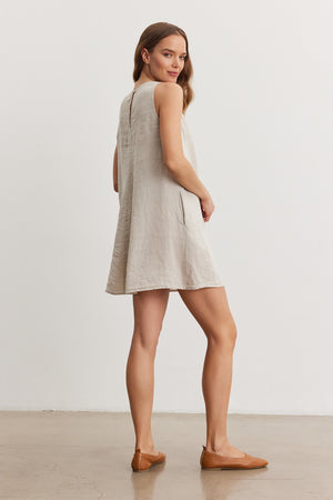 A woman in a Velvet by Graham & Spencer TIFFANY LINEN DRESS and brown loafers, standing in a studio facing away, slightly turning her head towards the camera.