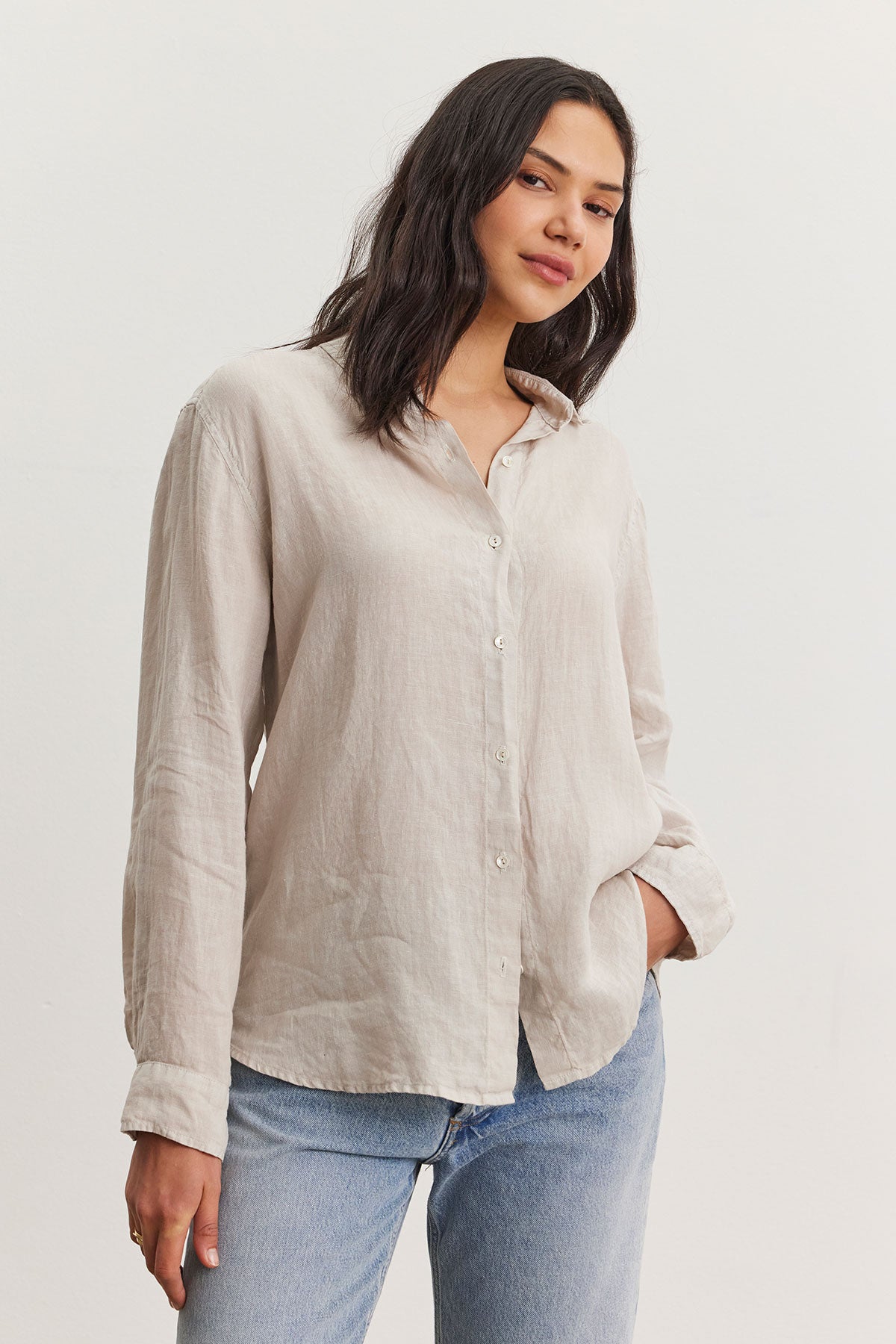   A woman wearing a Velvet by Graham & Spencer Willow Linen Button-Up Shirt paired with denim jeans stands confidently while slightly tilting her head, giving a relaxed pose in a bright indoor setting. 