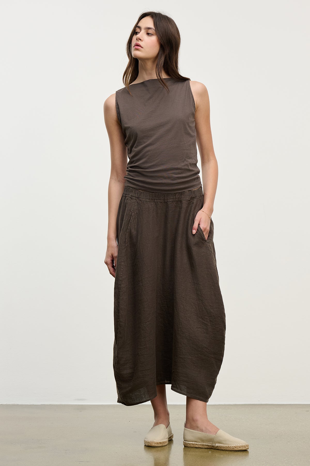   A woman stands against a plain white background wearing a sleeveless brown top, a Velvet by Graham & Spencer FAE LINEN SKIRT with an elastic waistband, and beige slip-on shoes. 