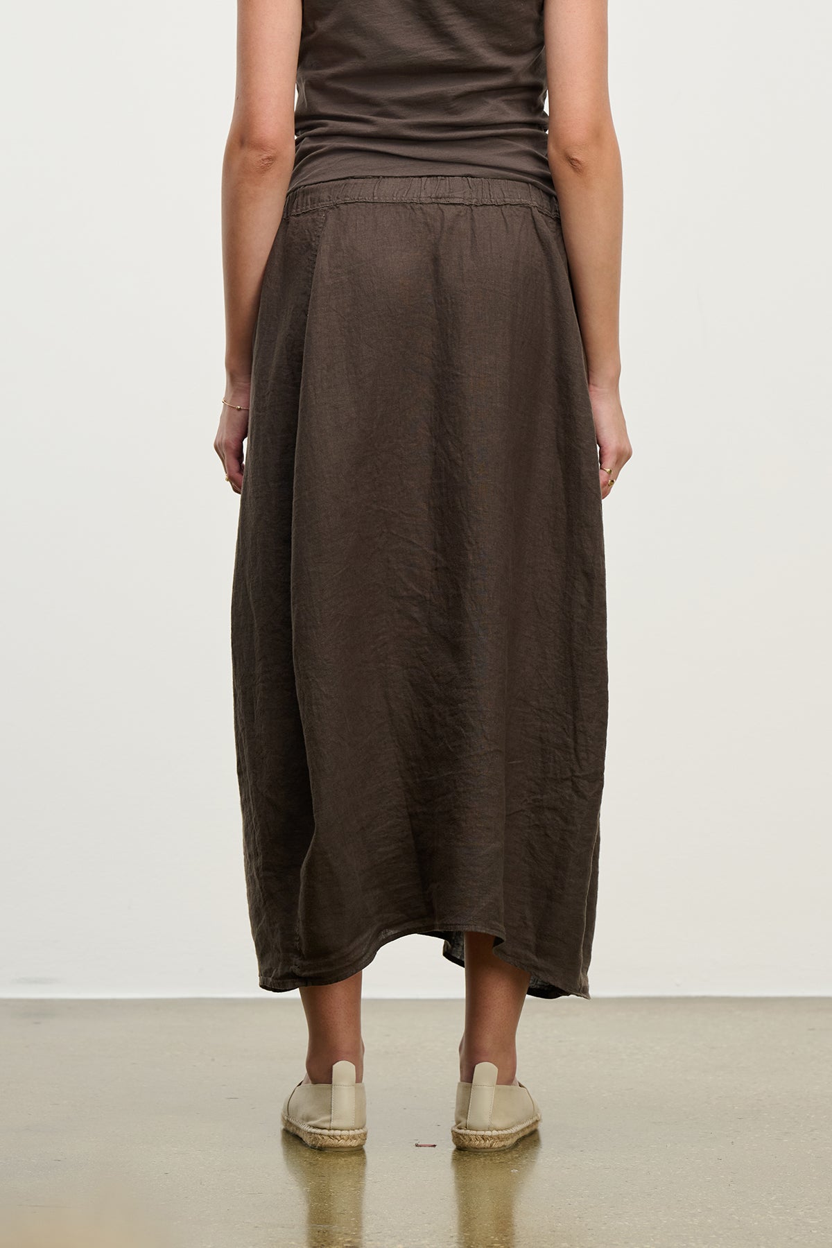   A person wearing a brown sleeveless top and the FAE LINEN SKIRT by Velvet by Graham & Spencer, matched with light slip-on shoes on a smooth floor, facing away. 