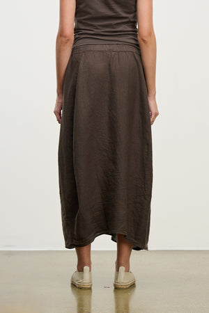 A person wearing a brown sleeveless top and the FAE LINEN SKIRT by Velvet by Graham & Spencer, matched with light slip-on shoes on a smooth floor, facing away.