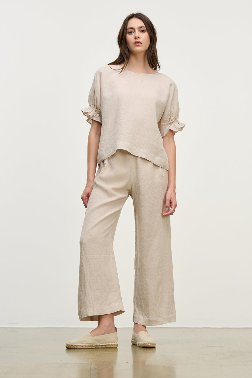 A woman models the Beige LOLA LINEN PANT by Velvet by Graham & Spencer. The outfit features a loose-fitting top and wide-leg, pull-on trousers with an elastic waist, set against a plain white background.