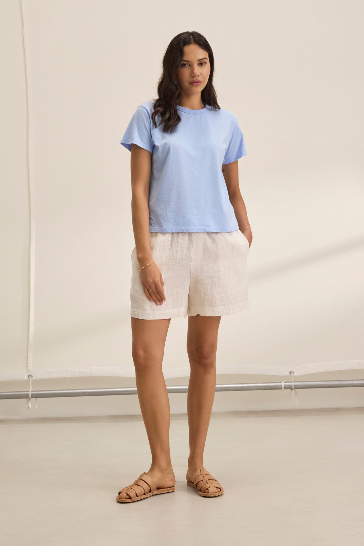   A woman in a light blue T-shirt, Velvet by Graham & Spencer's AMBER LINEN SHORT in beige with a drawstring elastic waist, and sandals stands in a minimalistic room. 