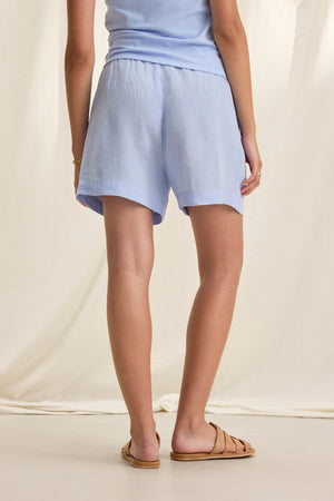 A person in a light blue sleeveless top and AMBER LINEN SHORTS by Velvet by Graham & Spencer, featuring a drawstring elastic waist, stands with their back turned against a cream backdrop.