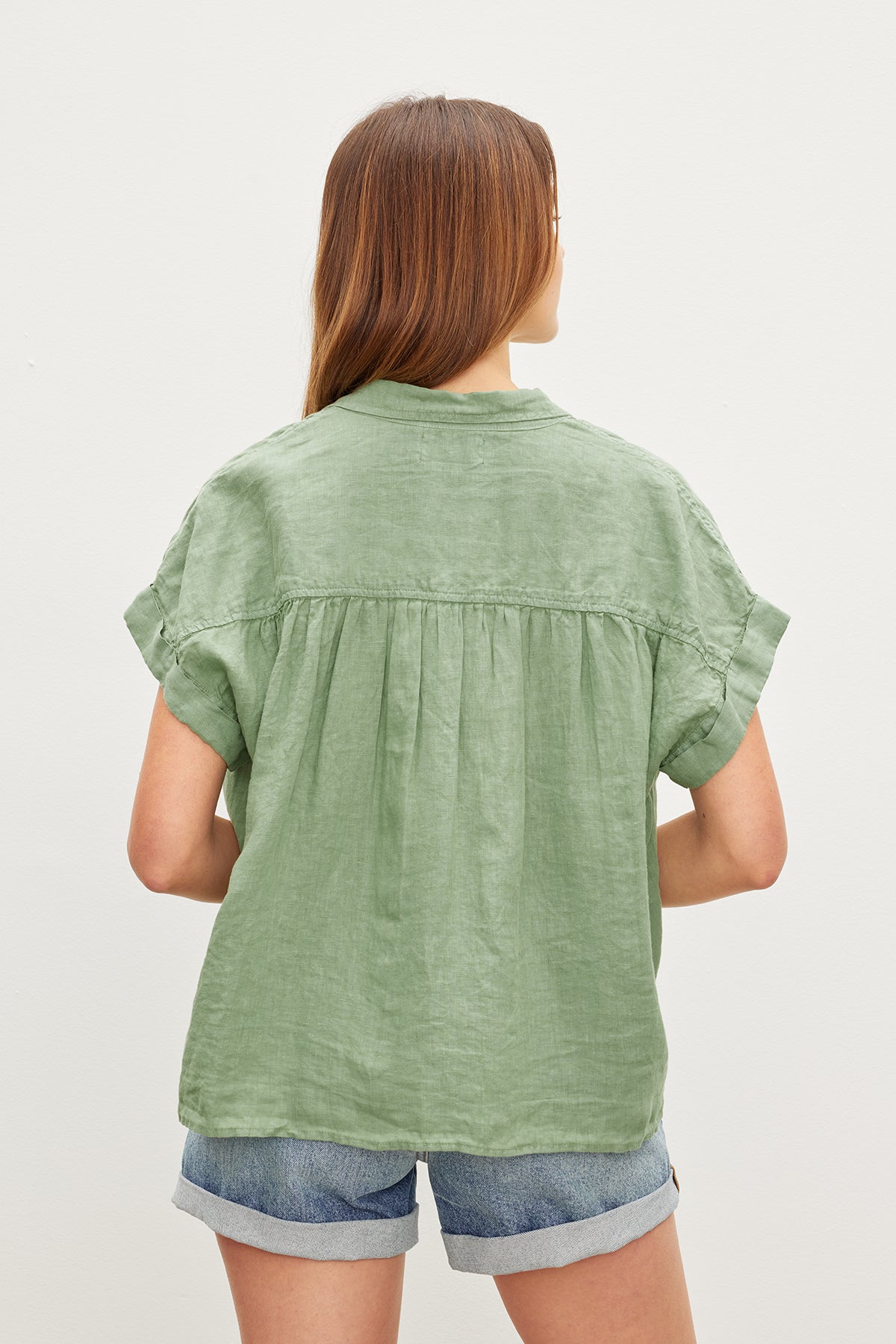   A person with long hair faces away, wearing the ARIA LINEN TOP by Velvet by Graham & Spencer, a green short-sleeve shirt with a classic collar, paired with denim shorts. 