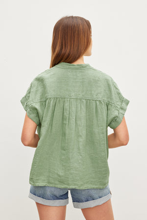 A person with long hair faces away, wearing the ARIA LINEN TOP by Velvet by Graham & Spencer, a green short-sleeve shirt with a classic collar, paired with denim shorts.