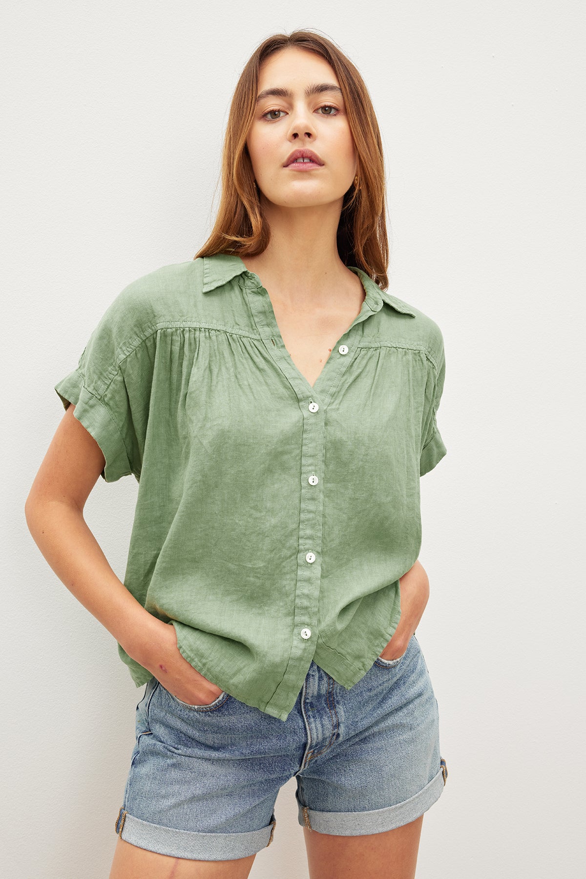   Against a white backdrop, a person wears the ARIA LINEN TOP by Velvet by Graham & Spencer, featuring a classic collar in green, paired with denim shorts and hands casually tucked into pockets. 