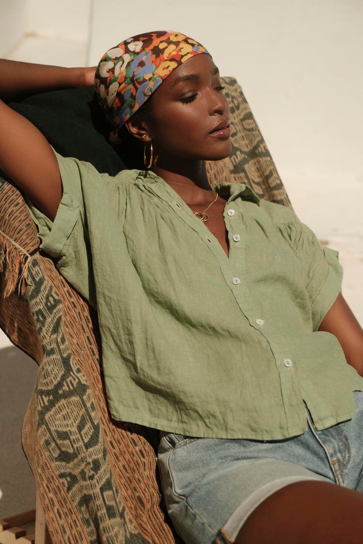   Someone in a Velvet by Graham & Spencer ARIA LINEN TOP, paired with denim shorts, sits comfortably on a patterned chair, sporting a colorful headscarf and gold hoop earrings. 