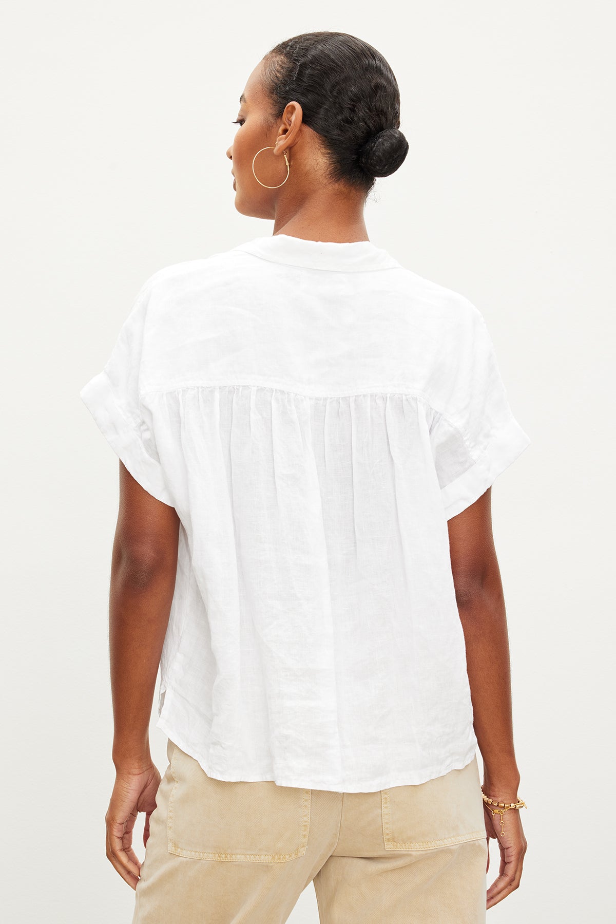   A person is seen from the back, wearing a white ARIA LINEN TOP by Velvet by Graham & Spencer with short sleeves and a classic collar, paired with beige pants. Their hair is tied back in a bun. 