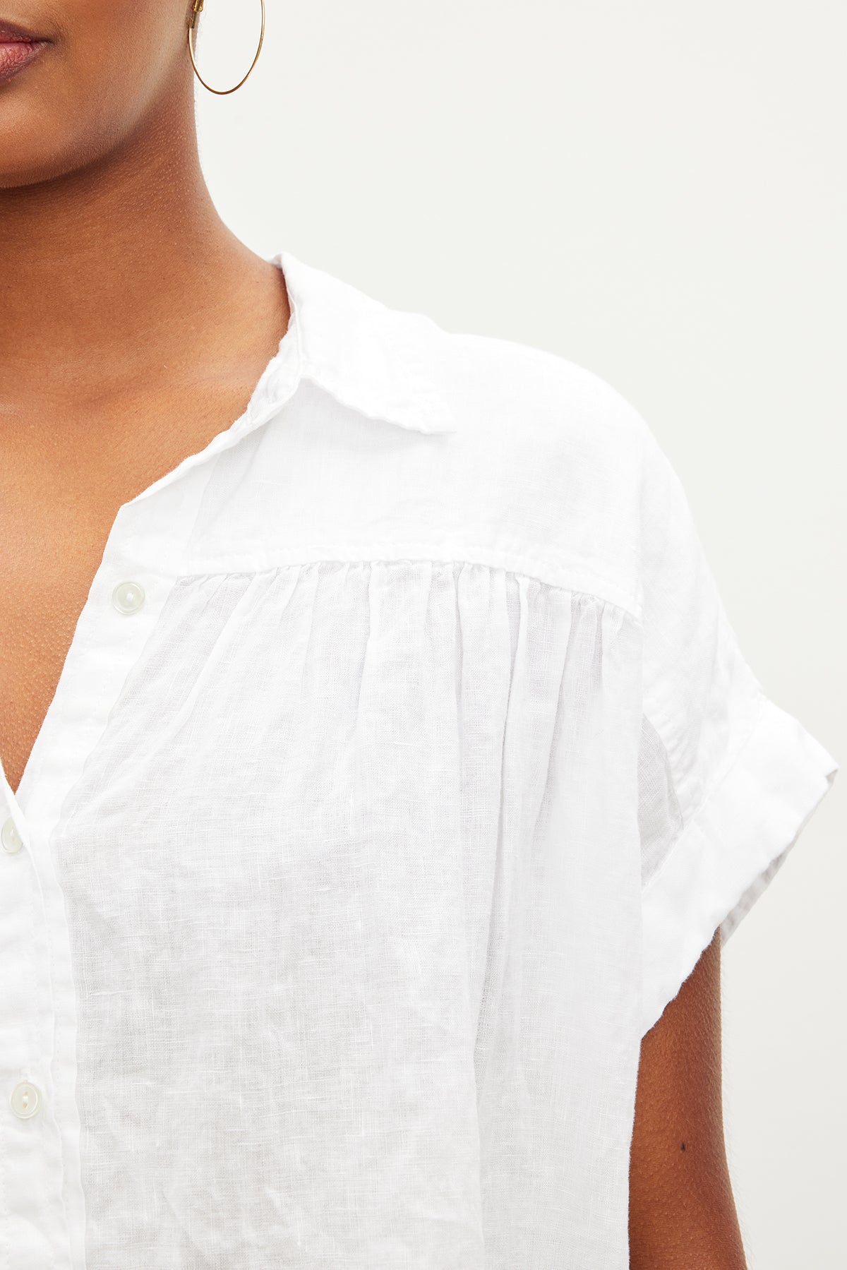   A person is wearing the ARIA LINEN TOP by Velvet by Graham & Spencer, a white collared short-sleeve shirt with pleated shoulder details. They are also seen with an earring. 