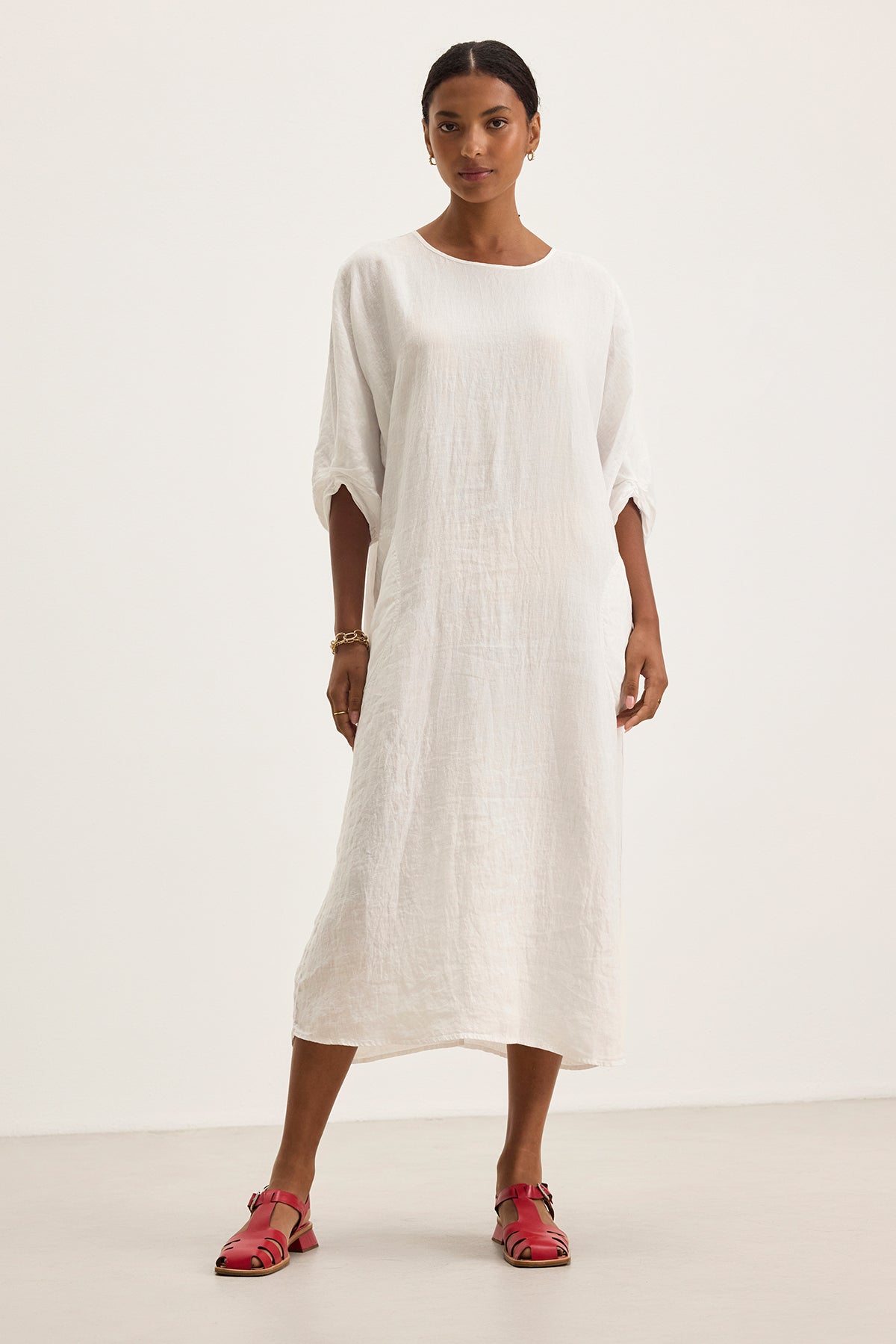 A person wearing the CARMEN LINEN DRESS by Velvet by Graham & Spencer, featuring a long white fabric and relaxed silhouette, pairs it with red sandals against a plain background.-38662648824001