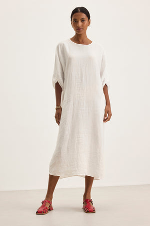 A person wearing the CARMEN LINEN DRESS by Velvet by Graham & Spencer, featuring a long white fabric and relaxed silhouette, pairs it with red sandals against a plain background.