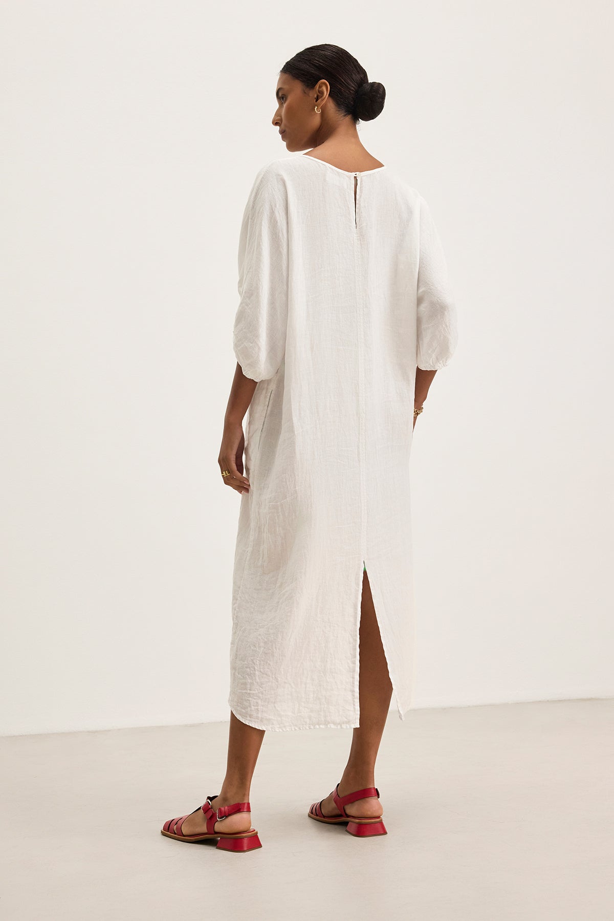   A person wearing the CARMEN LINEN DRESS by Velvet by Graham & Spencer, with a relaxed silhouette and back slit for elegant flow, pairs it with red sandals against a plain background. 