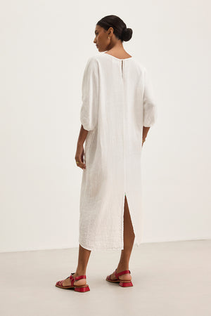 A person wearing the CARMEN LINEN DRESS by Velvet by Graham & Spencer, with a relaxed silhouette and back slit for elegant flow, pairs it with red sandals against a plain background.