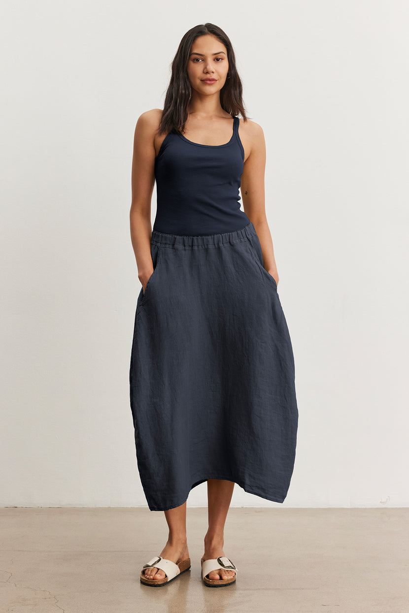 A woman wearing a navy tank top and the FAE LINEN SKIRT by Velvet by Graham & Spencer stands against a plain background with her hands casually in her pockets, paired with white sandals.