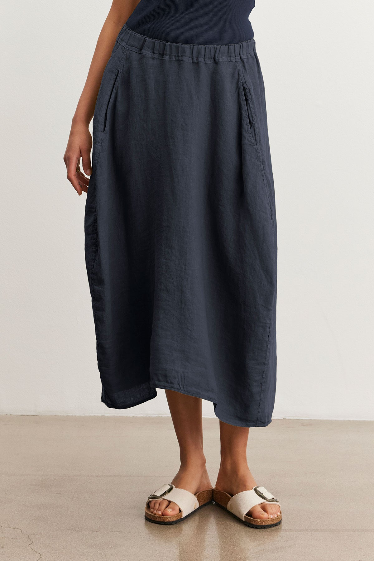   A person stands on a light brown floor, wearing beige sandals and a long, spring-weight dark gray FAE LINEN SKIRT with an elastic waistband by Velvet by Graham & Spencer. 