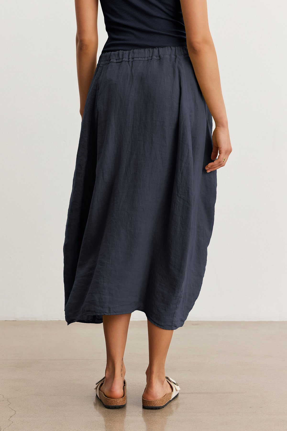 A barefoot person in a black shirt and long dark FAE LINEN SKIRT by Velvet by Graham & Spencer stands on a smooth floor, facing away.-38662669566145