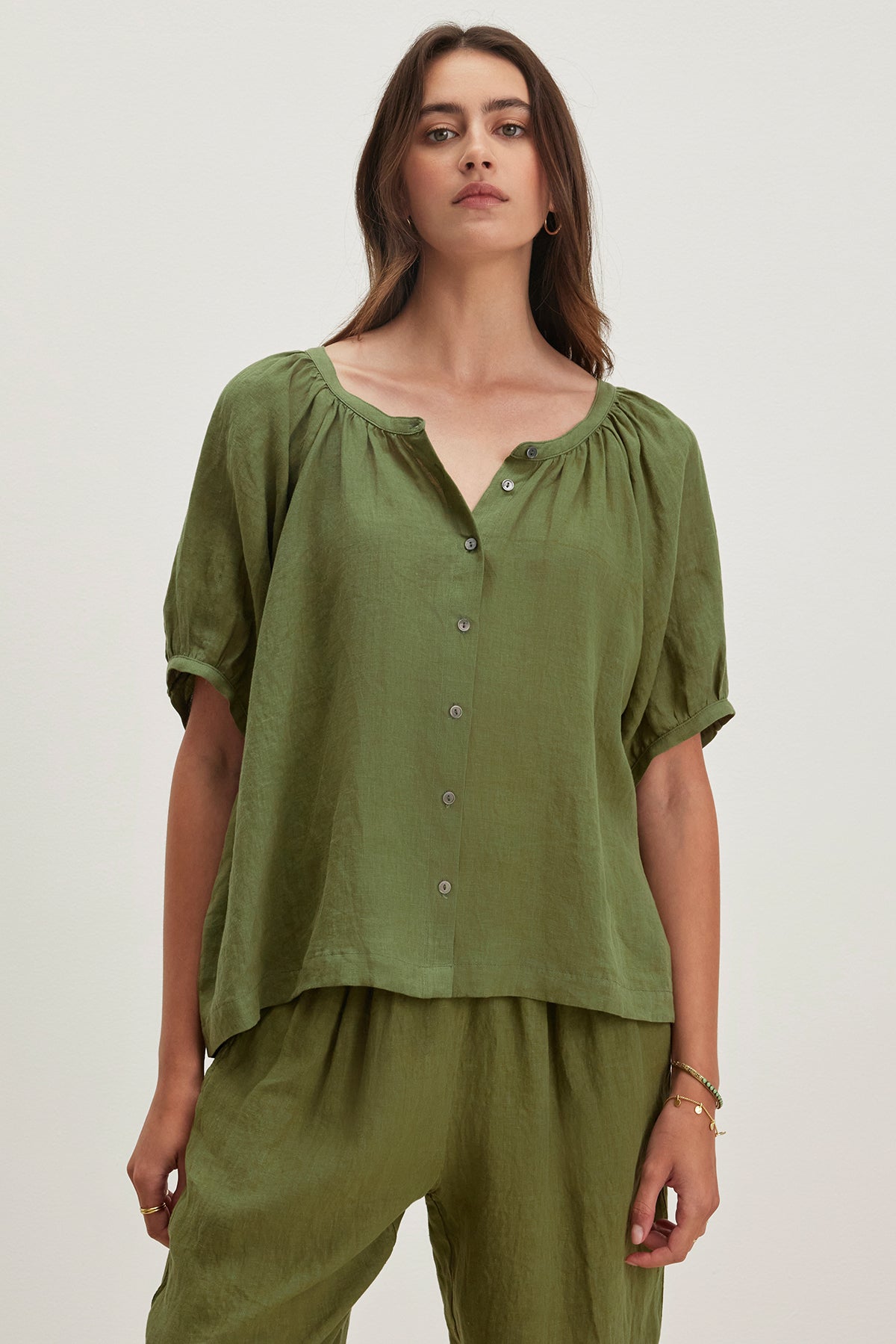   A woman elegantly poses against a plain backdrop, wearing the JAIMI LINEN TOP from Velvet by Graham & Spencer, a green button-down with puff sleeves, complemented by matching pants made from lightweight woven linen. 