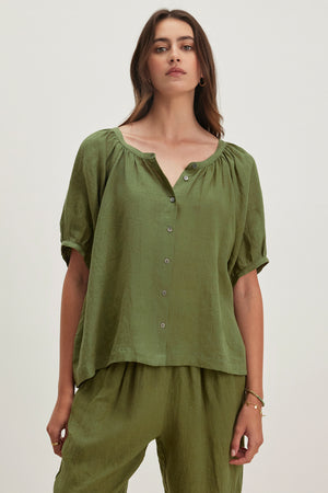 A woman elegantly poses against a plain backdrop, wearing the JAIMI LINEN TOP from Velvet by Graham & Spencer, a green button-down with puff sleeves, complemented by matching pants made from lightweight woven linen.