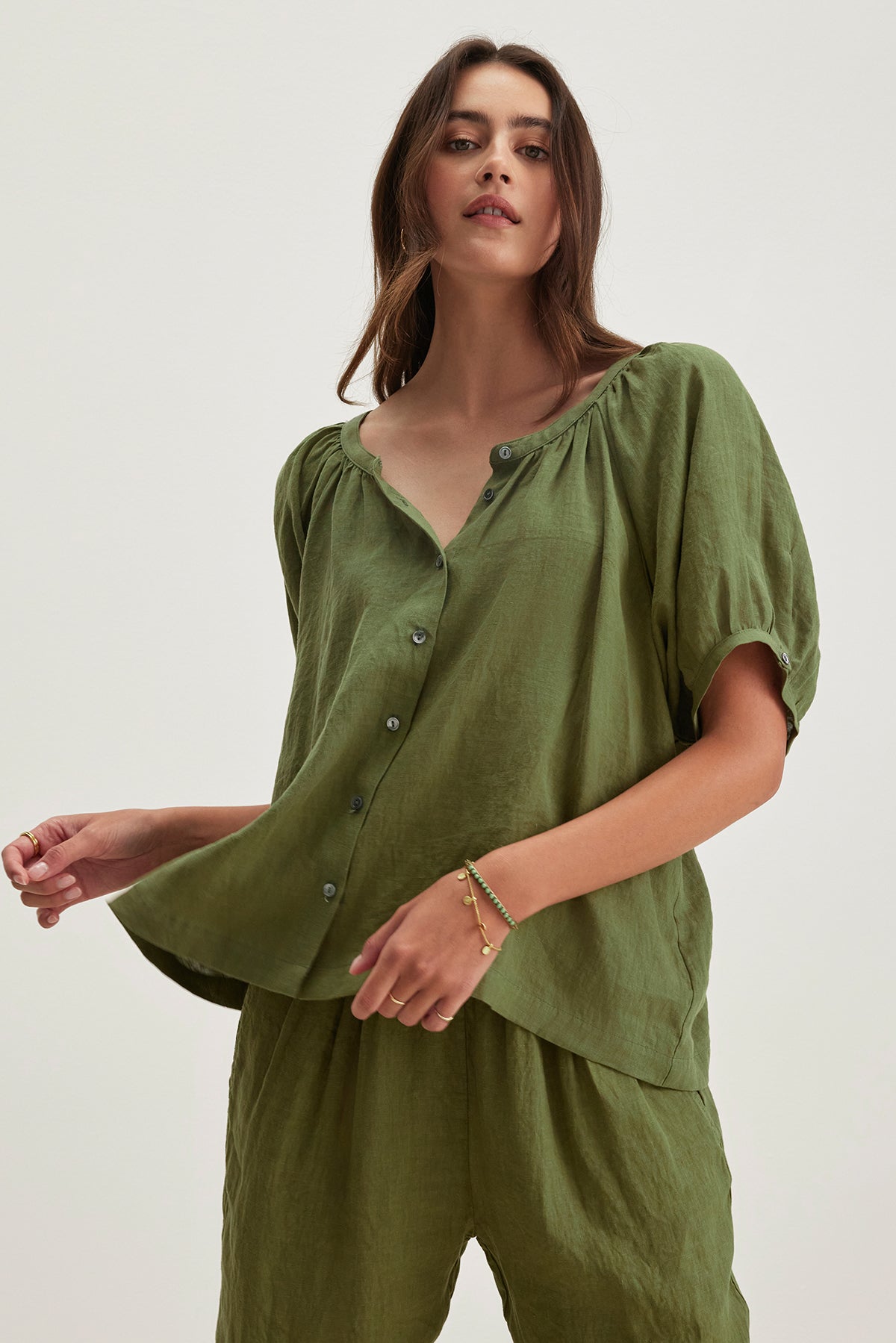   Someone wearing the JAIMI LINEN TOP from Velvet by Graham & Spencer, a loose-fitting green button-front blouse with puff sleeves and matching pants, stands against a light background, highlighting the lightweight woven linen fabric. 