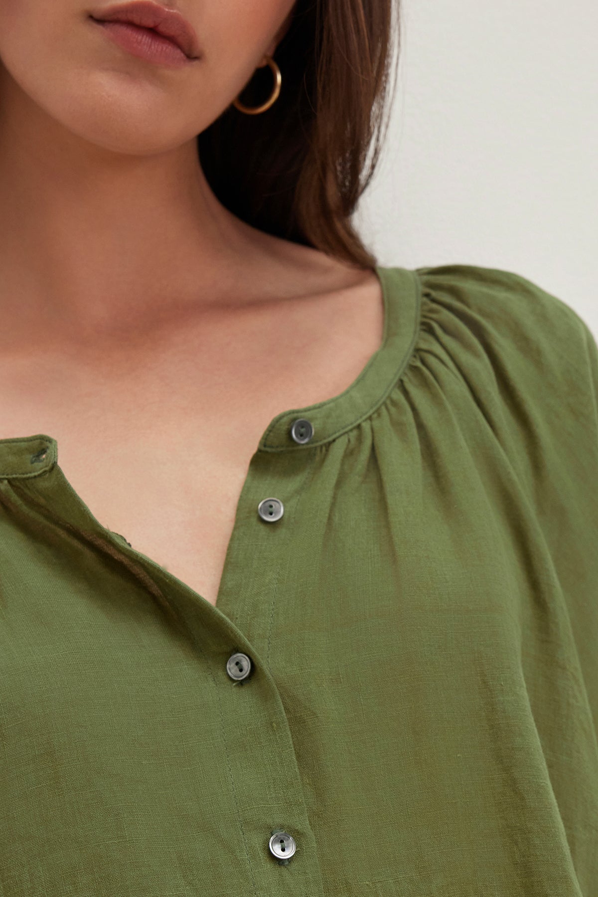   A person with long brown hair and an earring is wearing the JAIMI LINEN TOP by Velvet by Graham & Spencer, a green lightweight woven linen button-up with a gathered neckline. 