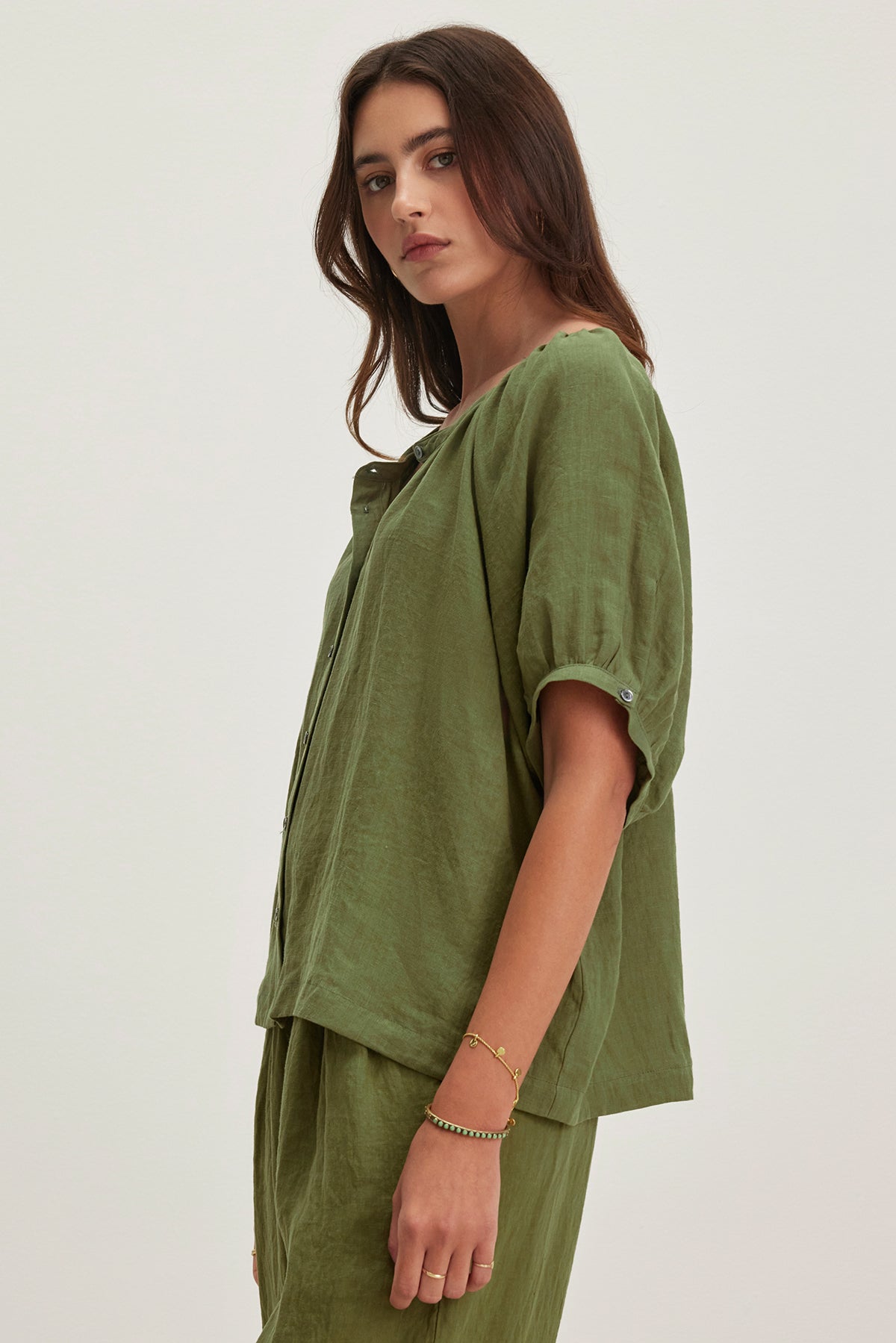   A person stands against a plain background, wearing the JAIMI LINEN TOP by Velvet by Graham & Spencer—a loose-fitting green blouse with puff sleeves and matching pants. The lightweight woven linen fabric adds airy elegance as they look to the side. 