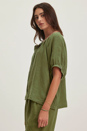 A person stands against a plain background, wearing the JAIMI LINEN TOP by Velvet by Graham & Spencer—a loose-fitting green blouse with puff sleeves and matching pants. The lightweight woven linen fabric adds airy elegance as they look to the side.