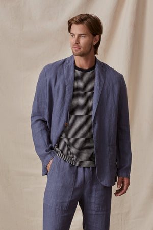 A man in a tailored fit, Blue Joshua Linen Blazer by Velvet by Graham & Spencer and striped t-shirt stands confidently against a neutral backdrop.