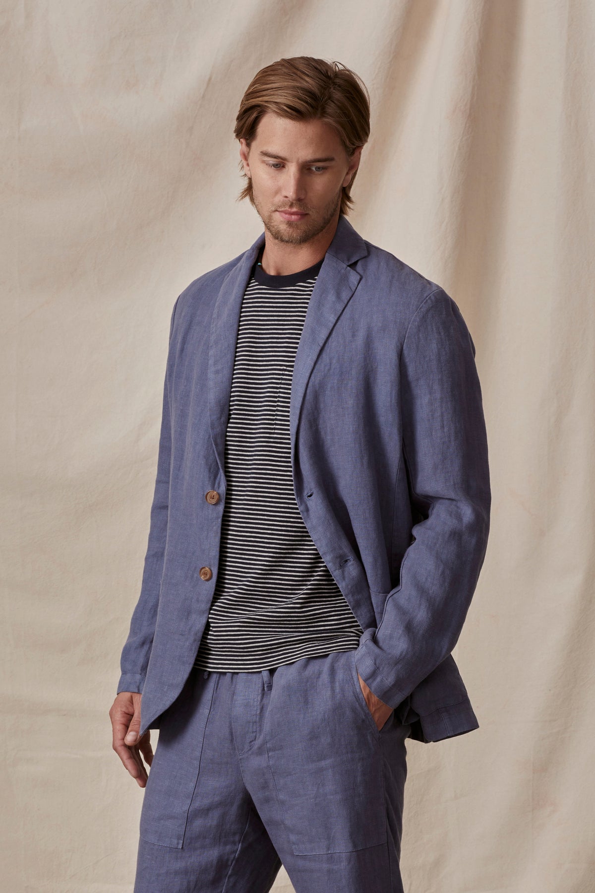   A man in a stylish blue Velvet by Graham & Spencer Joshua linen blazer and striped shirt, standing with hands in pockets against a neutral backdrop. 