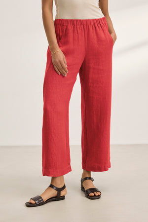 A person is indoors on a light floor, wearing Velvet by Graham & Spencer's stylish LOLA LINEN PANT in red with an elastic waist and paired with black sandals. Only the lower body is visible.