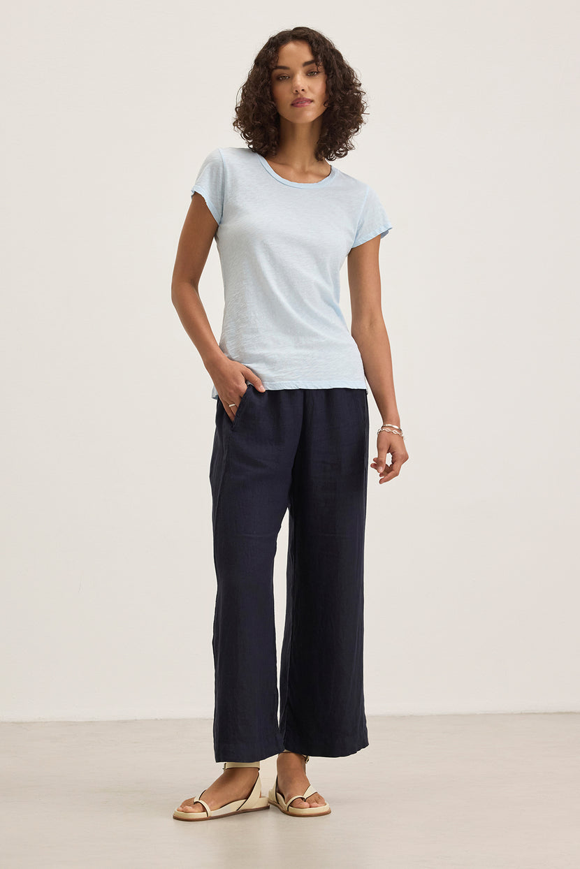 A person is wearing a light blue T-shirt, the LOLA LINEN PANT from Velvet by Graham & Spencer in dark wide-legged style with an elastic waist, paired with sandals against a plain background.