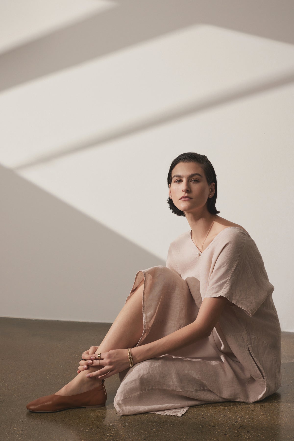   A woman in a Velvet by Jenny Graham Montana Linen Dress sits on the floor, leaning against a wall with diagonal light stripes, looking contemplatively towards the camera. 