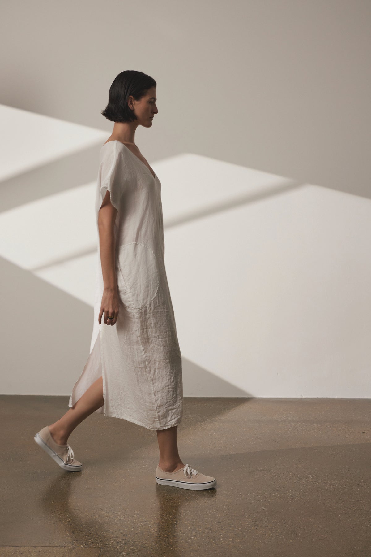   A person with short dark hair in a loose-fitting MONTANA LINEN DRESS by Velvet by Jenny Graham is walking in a well-lit room with a minimalist design. They are wearing white shoes and the background features shadows from window blinds, adding depth to the airy ambiance. 