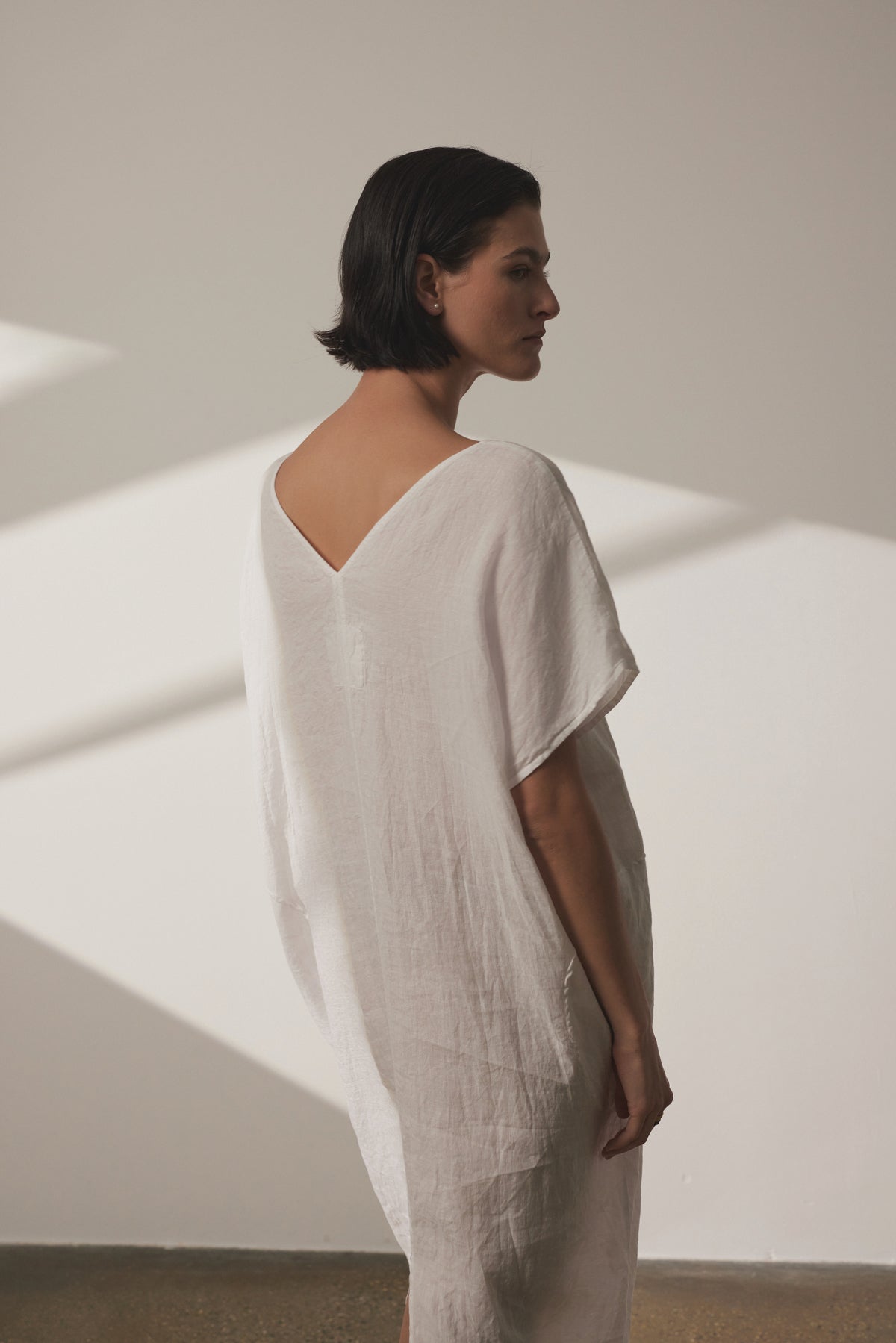   A person with short dark hair stands wearing a loose-fitting white MONTANA LINEN DRESS by Velvet by Jenny Graham, looking to the side in a well-lit room. 