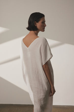 A person with short dark hair stands wearing a loose-fitting white MONTANA LINEN DRESS by Velvet by Jenny Graham, looking to the side in a well-lit room.