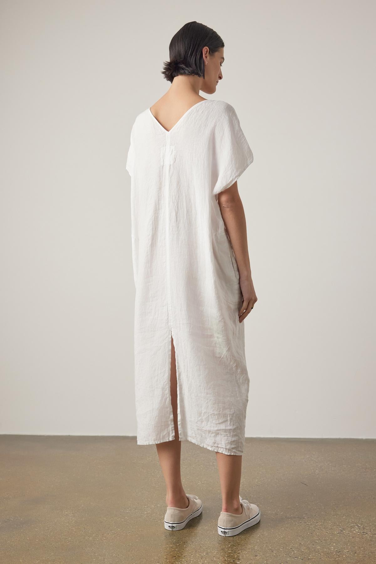   A person stands facing away, wearing a loose, knee-length MONTANA LINEN DRESS by Velvet by Jenny Graham with short sleeves and a low-cut back. They have short dark hair and are wearing white sneakers. The background is a plain, light-colored wall. 