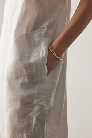 A close-up of a person wearing a light-colored, textured MONTANA LINEN DRESS by Velvet by Jenny Graham with their hand in the pocket. The person has a gold bracelet on their wrist.