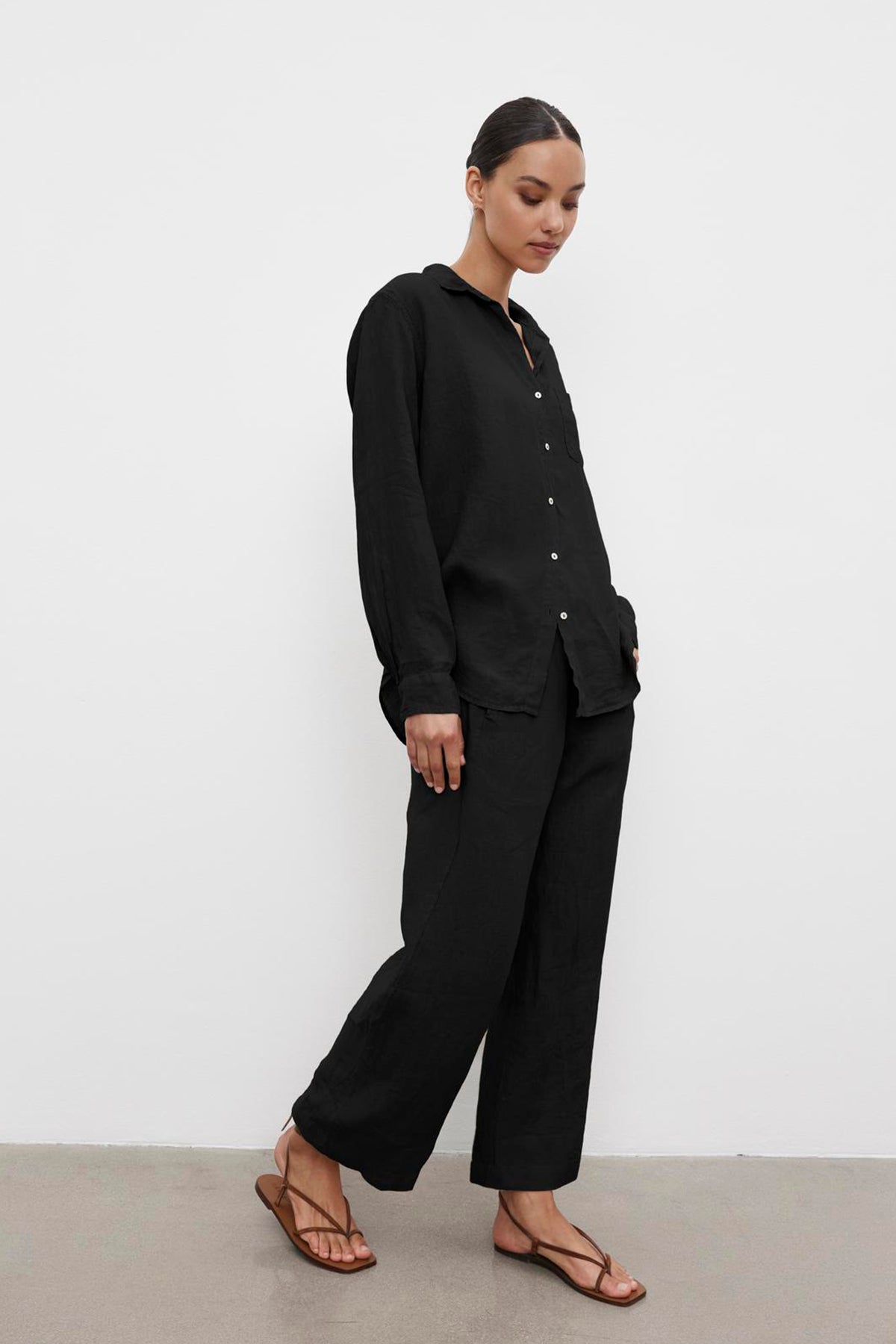   A person stands against a white wall, dressed in a black MULHOLLAND LINEN SHIRT by Velvet by Jenny Graham with a relaxed silhouette and black pants, wearing brown sandals. They have their hands in their pockets and are looking down. 