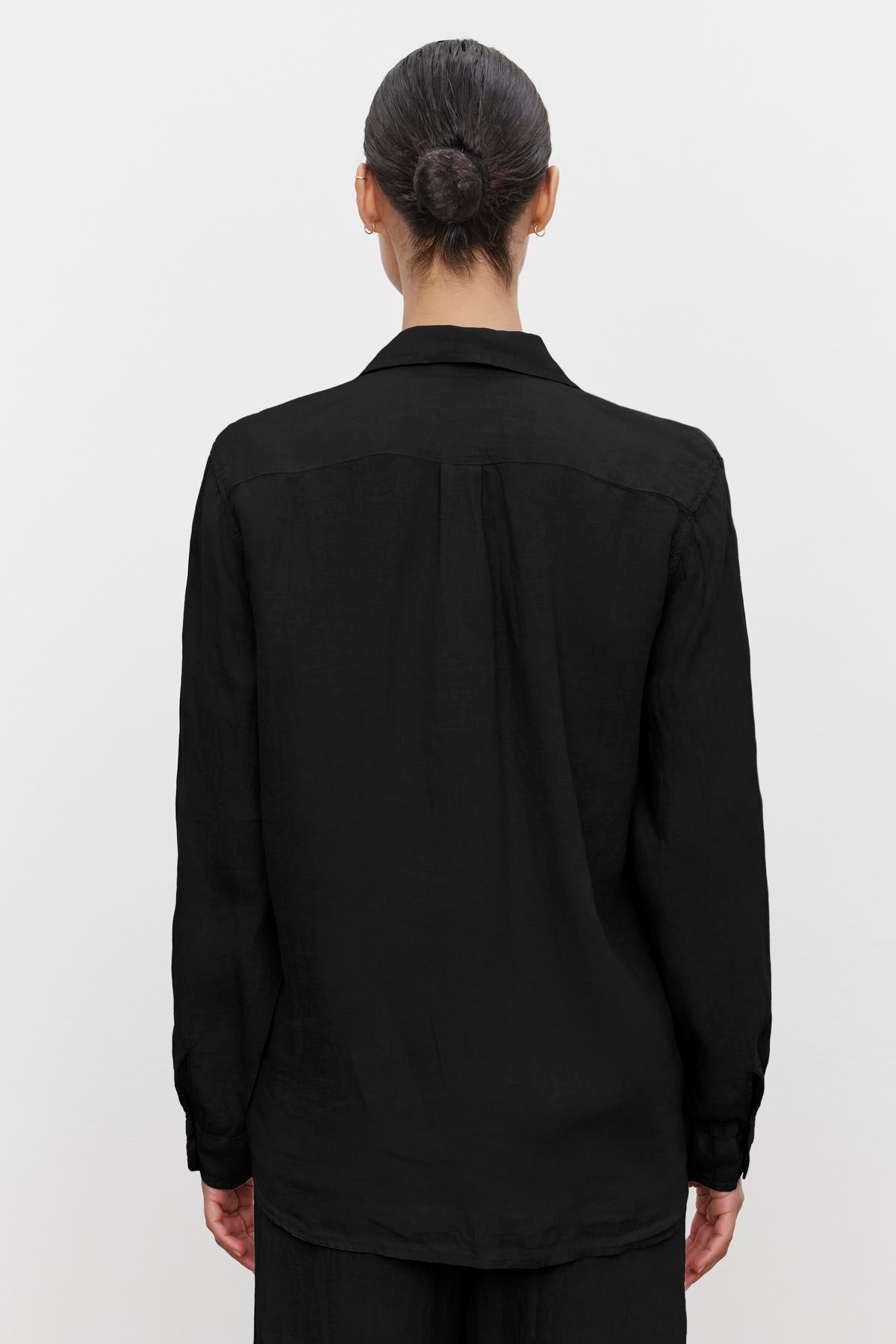 A person with dark hair in a bun is wearing a long-sleeved black Velvet by Jenny Graham MULHOLLAND LINEN SHIRT with a relaxed silhouette and scooped hemline, facing away from the camera, showing the back view of the shirt.-36463599616193