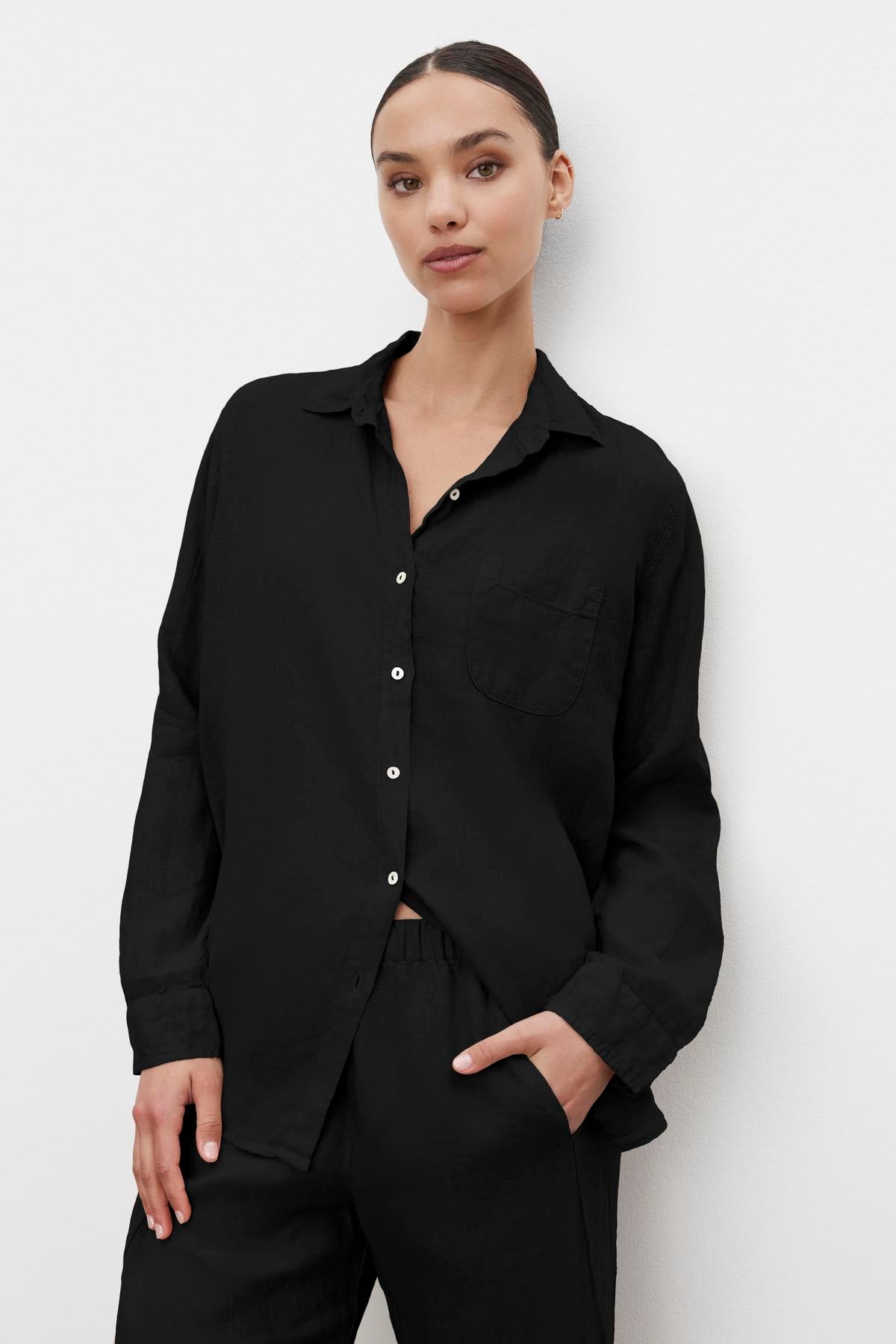 Woman wearing a black MULHOLLAND LINEN SHIRT by Velvet by Jenny Graham and black pants, standing against a plain white background with one hand in pocket.-36463599517889