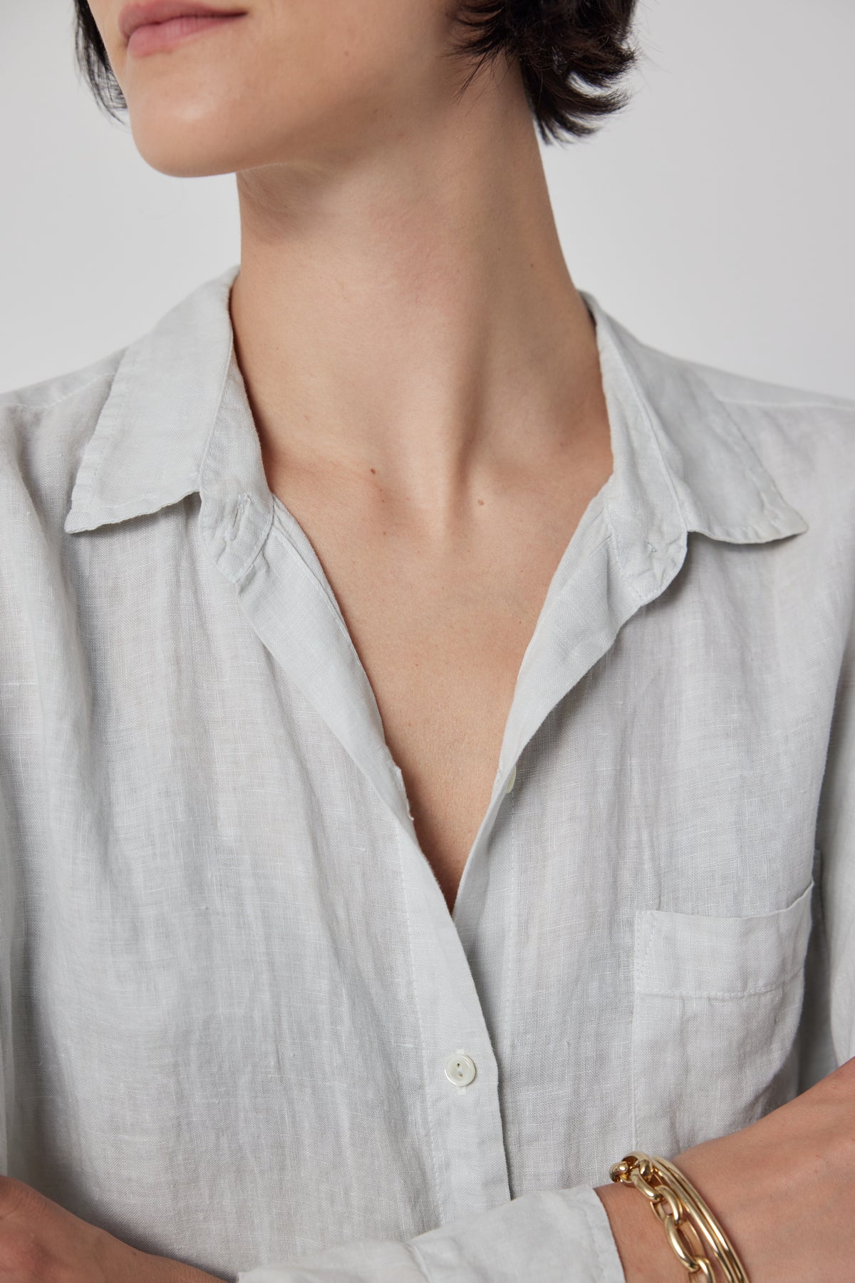 Close-up of a person wearing the Velvet by Jenny Graham MULHOLLAND LINEN SHIRT with a chest pocket. The person has short, dark hair and is accessorizing with a gold bracelet. The relaxed silhouette of the shirt features a scooped hemline. The background is plain white.-36212529692865