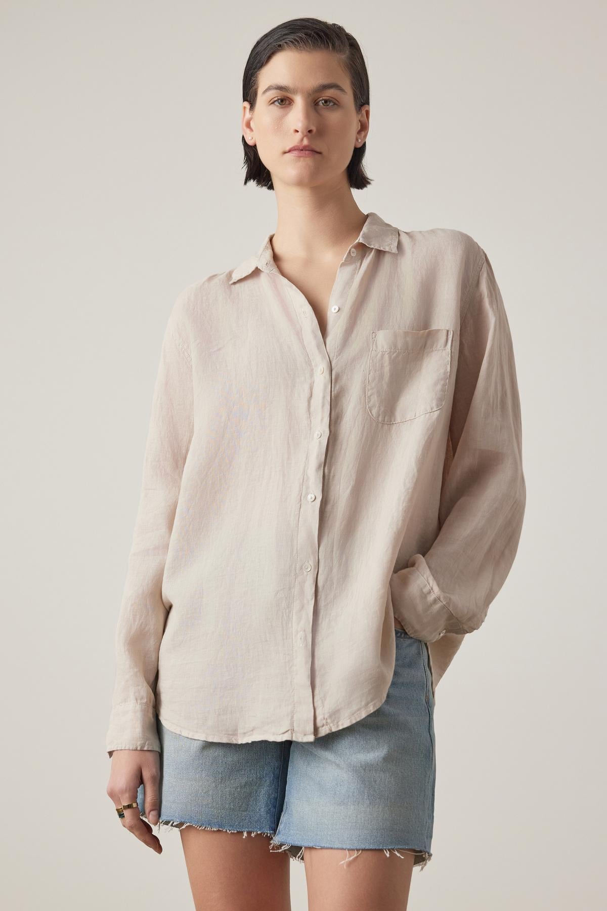 Person with short dark hair wearing a Velvet by Jenny Graham MULHOLLAND LINEN SHIRT featuring a relaxed silhouette and scooped hemline, paired with light blue denim shorts, stands against a plain background.-36890595360961